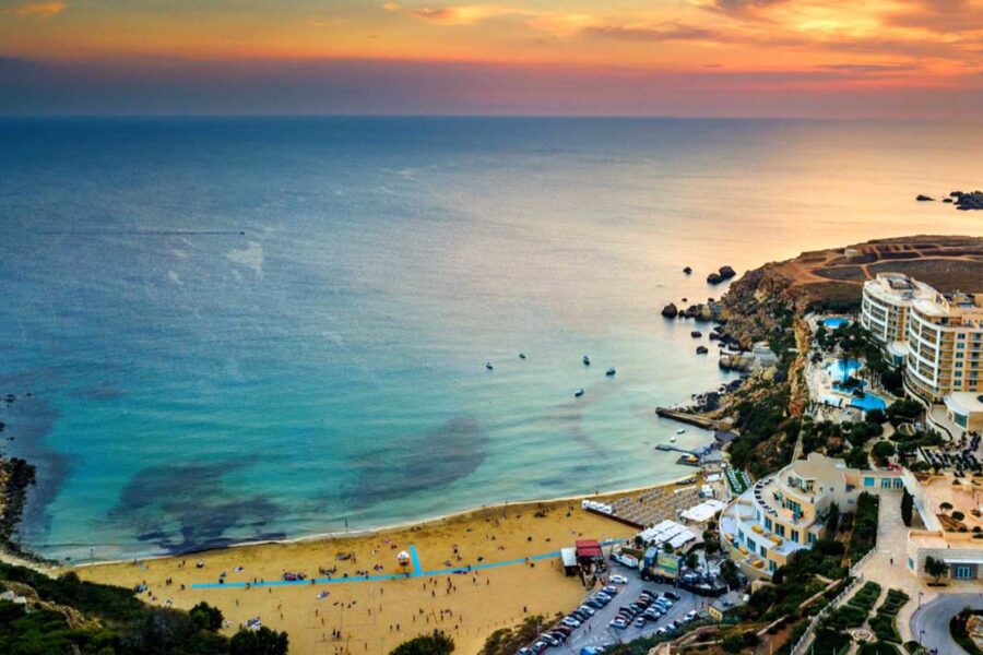 Aerial drone view of the golden sandy beach, cliffside hotel, and azure coastal waters at Ir-Ramla tal-Mixquqa (Golden Bay) in Malta.