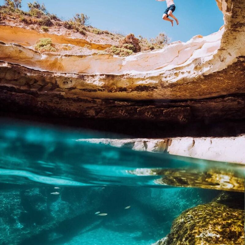 A man mid-air leaping into the deep, crystal-clear turquoise waters of St.Peter's Pool natural pool in southern Malta during a private boat tour.